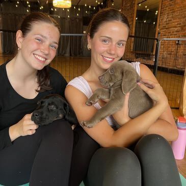 Two smiling women sitting with puppies in a cozy indoor space.

