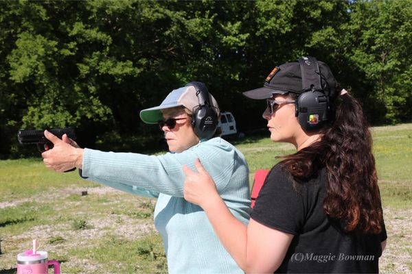 Two women at an outdoor shooting range, one aiming a handgun, the other instructing.