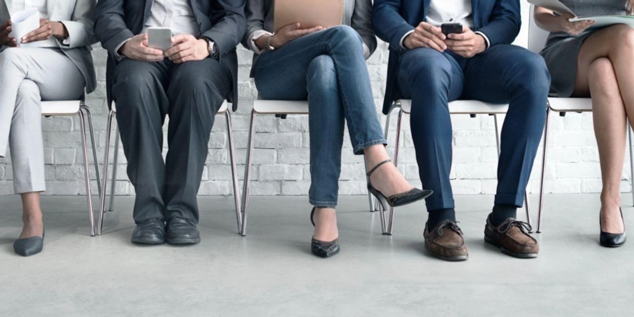 People holding cell phones and folders sitting in a line of chairs waiting for job interviews. 