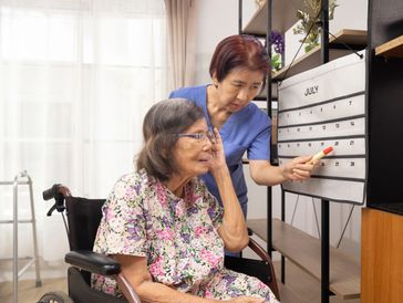 Caregiver assists elderly woman in wheelchair by pointing to dates on large wall calendar.