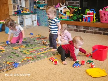 Four toddlers playing with toy cars on a carpeted floor in a colorful playroom.