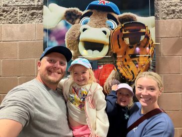 Family smiling in front of a mascot poster at a baseball stadium.