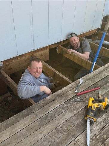 Two men working under a wooden deck, smiling.