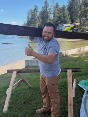 Man holding a wooden plank by a lakeside, giving thumbs up.