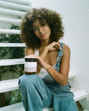 Woman with curly hair holding a jar of curl mask sitting on white stairs.