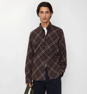 Man wearing a brown plaid shirt and dark pants, holding a green bag strap.