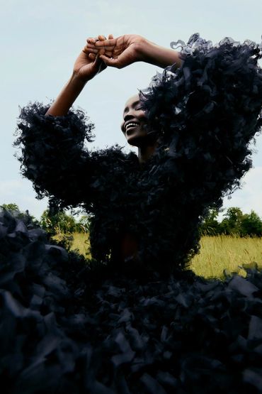 A joyful person in a voluminous black ruffled outfit poses outdoors with arms raised.