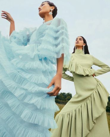 Two women pose in elegant ruffled dresses outdoors, showcasing high fashion style.
