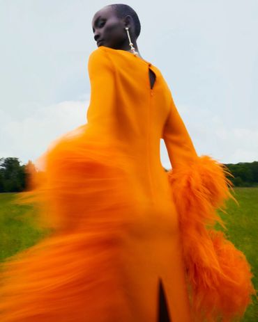 Model in vibrant orange dress with feathered sleeves in a blurred motion outdoors.