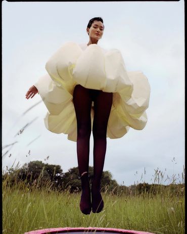 Person in a voluminous cream dress and black tights jumping on a trampoline in a grassy field.