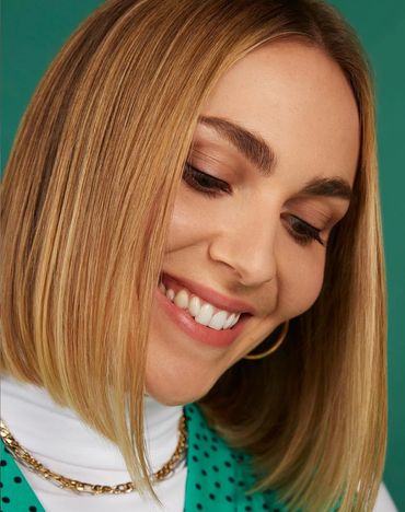 Close-up of a smiling woman with straight blonde hair and gold accessories.