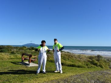 Two men in white overalls and neon hoodies stand on a grassy area by the beach.