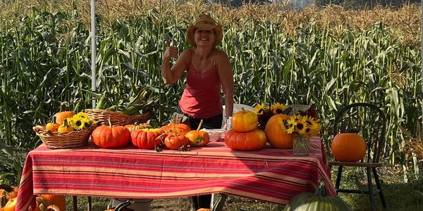 Woman selling pumpkins and sunflowers at a farm stand by a cornfield.