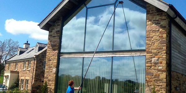 Person cleaning large glass windows of a stone house with a long pole.
