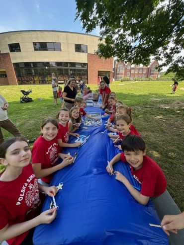 Children in red shirts sitting around a long blue table outdoors, smiling and crafting.