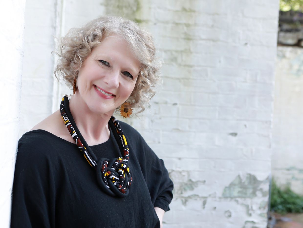 Smiling woman with curly hair and colorful necklace against a white brick wall.