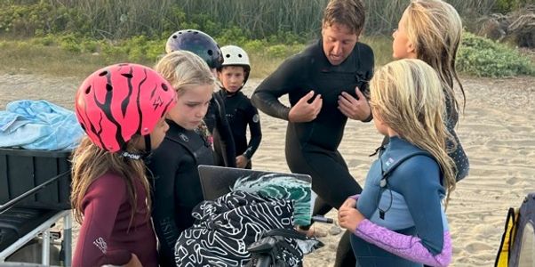 Children in wetsuits and helmets at the beach.