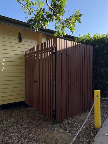 Brown wooden fence near yellow building with green foliage.