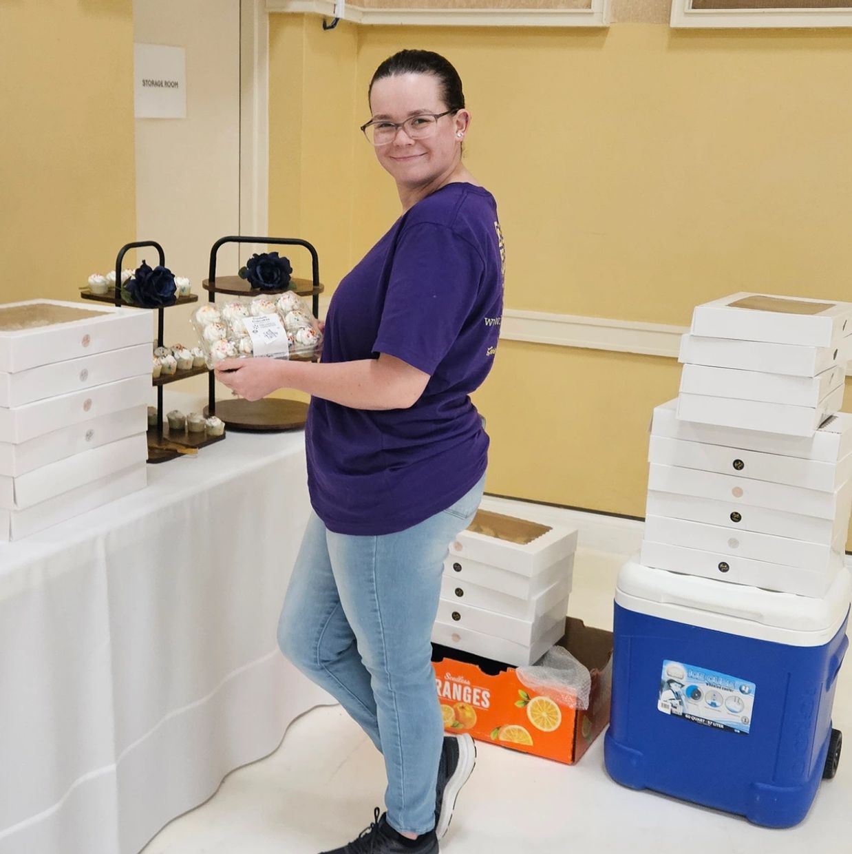 A woman in a purple shirt holding a pack of cupcakes, smiling at the camera.