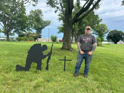 Man stands next to a military silhouette and cross in a grassy field.