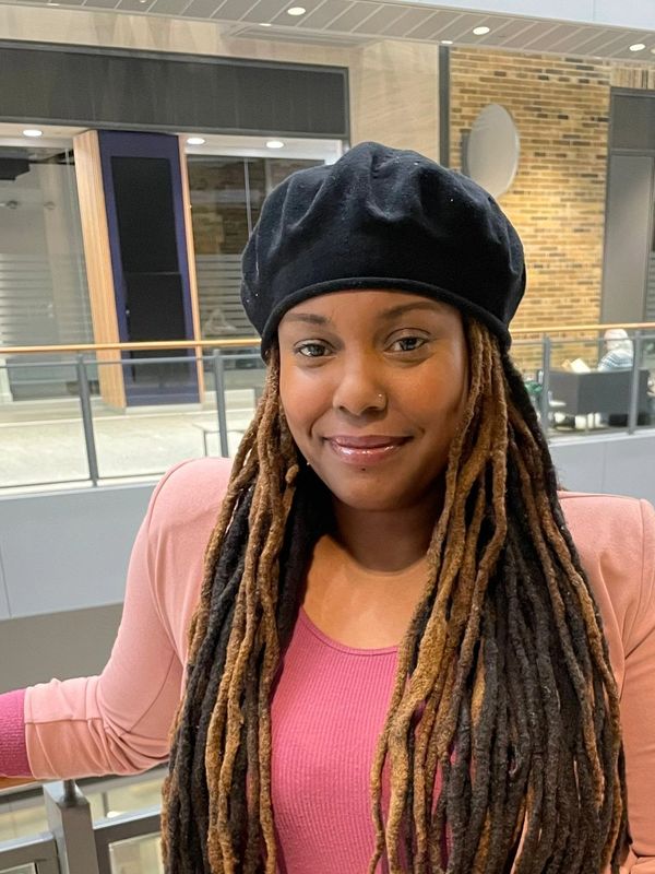 Smiling woman with long dreadlocks wearing a black beret and pink outfit indoors.