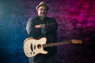 Man with a guitar, wearing a hat and glasses, smiling confidently.