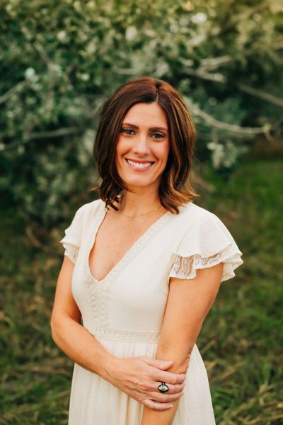 Smiling woman in a white dress stands outdoors with greenery behind her.