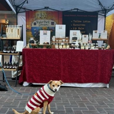 A dog in a striped sweater sits in front of a handmade product stall at a market.