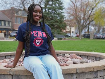 Smiling young woman wearing a Penn shirt sitting on a stone ledge outdoors.