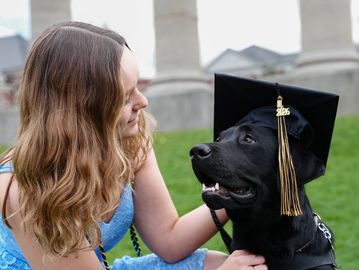A woman admires a black dog wearing a graduation cap outdoors.