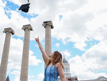Graduate tossing cap in the air by historic columns on a sunny day.