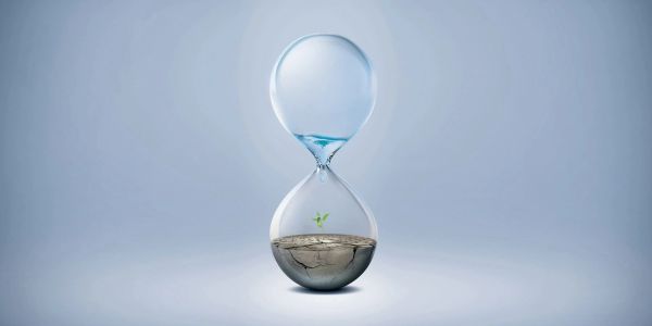 Hourglass with water and small plant growing in cracked soil.