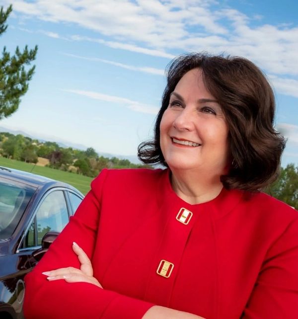 Smiling woman in a red jacket standing outdoors with a car and trees in the background.