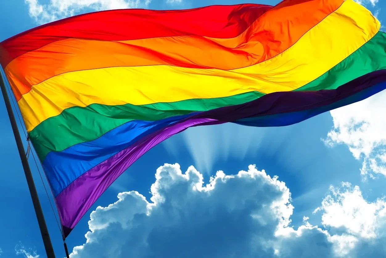 A rainbow flag flying against a blue sky with clouds
