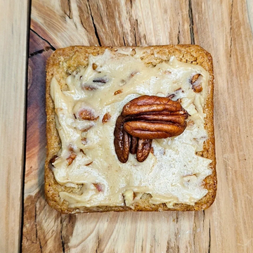 Square cookie with creamy frosting topped with pecans on a wooden surface.