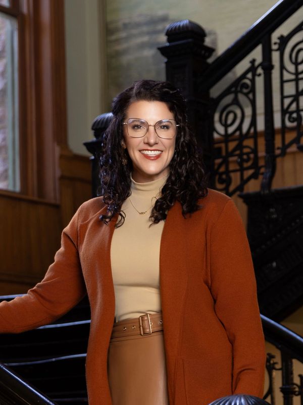 Woman in glasses and brown outfit smiling on ornate staircase indoors.