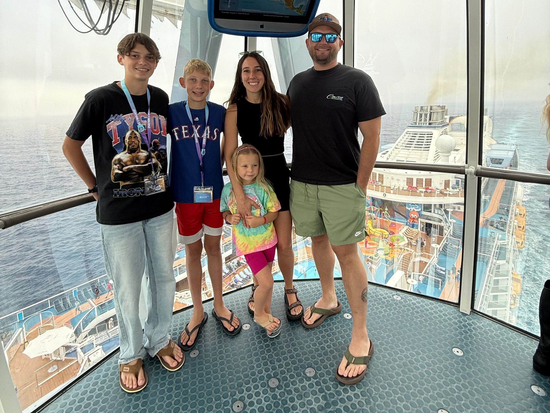 Family of five posing inside a glass gondola on a cruise ship under cloudy skies.