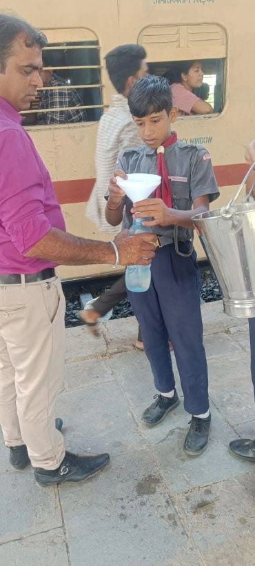 Boy in scout uniform helping man fill water bottle at train station.