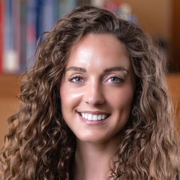 Smiling woman with long curly hair in a softly blurred indoor setting.