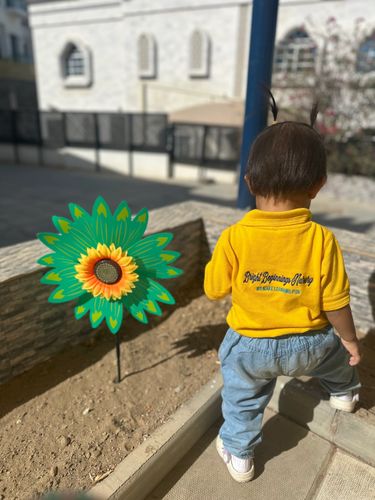 A toddler in a yellow shirt stands near a colorful flower decoration outdoors at Bright Beginnings Nursery. 