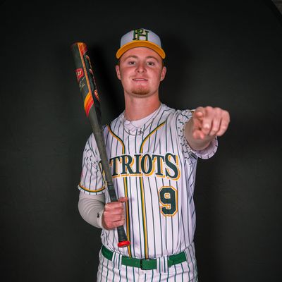 Baseball player in Patriots uniform holding bat and pointing forward.