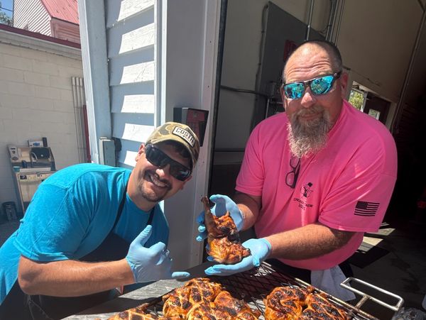 Two men presenting grilled chicken at an outdoor cookout.