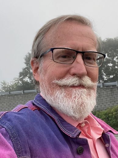 Smiling man with glasses and white beard wearing a purple jacket outdoors.