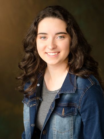Young woman with curly hair smiling, wearing a denim jacket and gray shirt.