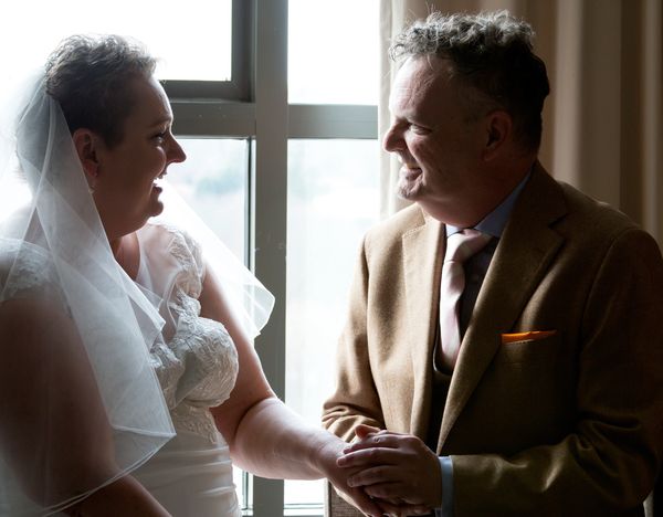 Bride and groom smiling at each other, holding hands by a window.