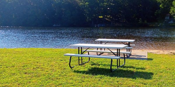 Three picnic tables on grass by a calm lake with trees in the background.