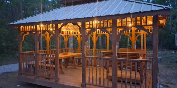 Wooden gazebo illuminated with warm lights in a forest setting at dusk.