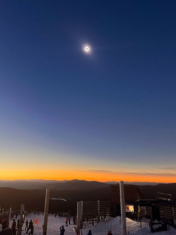 Total solar eclipse over a snowy mountain at sunset with people watching.