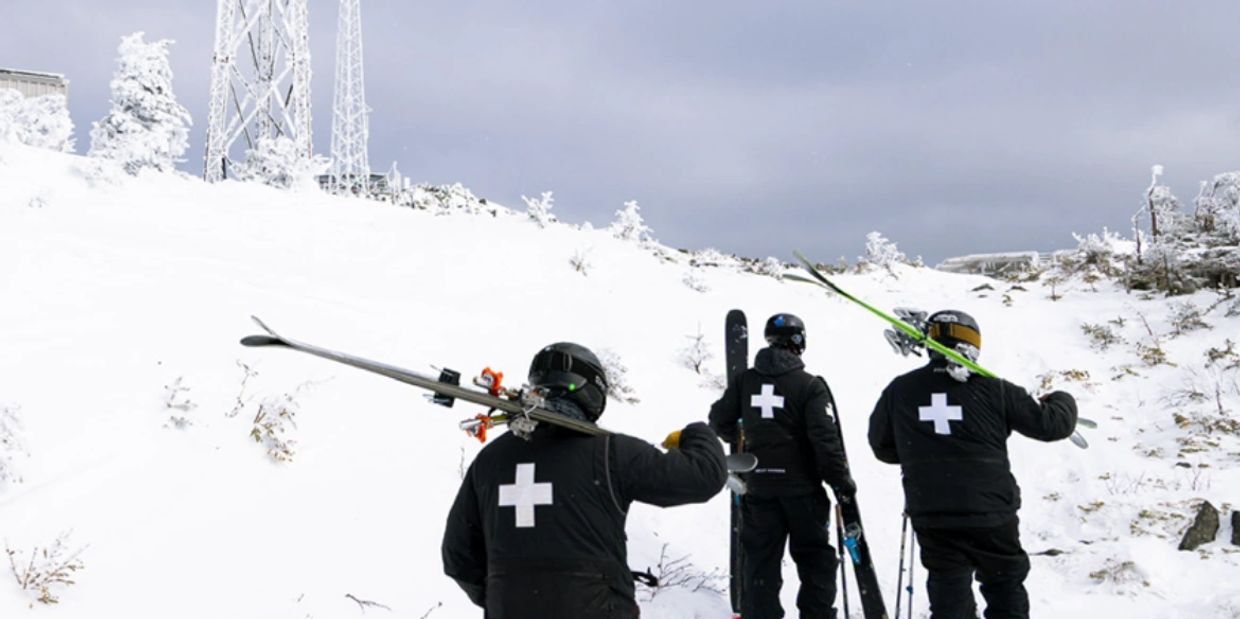 Three ski patrollers walk up a snowy hill carrying skis.
