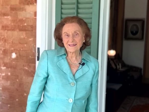 Elderly woman in a turquoise suit smiling by a doorway.